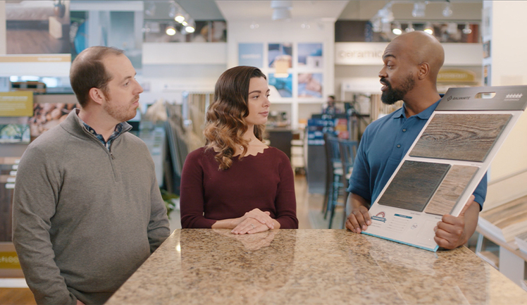 A man and woman talk with a carpet store employee as they go over different carpet samples. The employee is holding up a board of different samples.